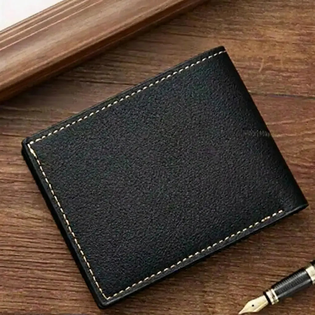 Back view of the closed Arlen black bifold wallet resting on a dark wood table next to a fountain pen, highlighting its slim profile and textured leather finish.